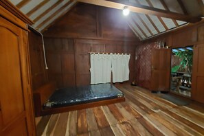 A room with a wooden floor, a bed with a mattress, a window with curtains, and a door leading to another room. - Mbok Niem Cafe and Cottage (Bangunjiwo)