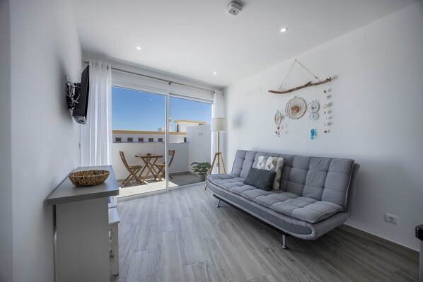 Ein modernes Wohnzimmer mit grauem Sofa, einem flachbildfernseher an der Wand, einem Esstisch mit Stühlen und einer großen Glasschiebetür, die auf einen Balkon im Freien führt.