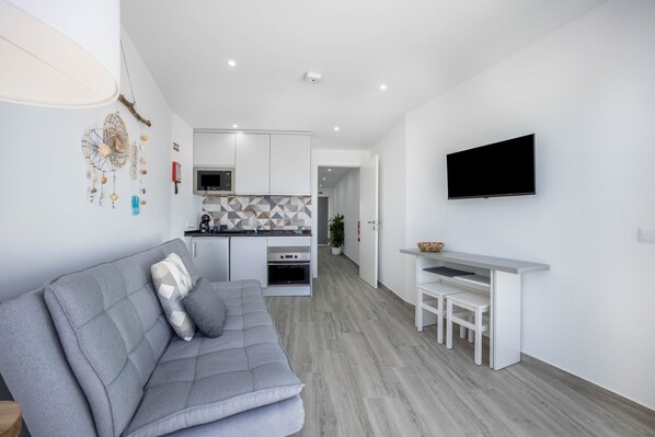 Un appartement moderne avec un canapé gris, une cuisine dotée de meubles blancs, un téléviseur fixé au mur et un petit coin repas.