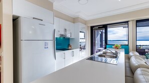 A modern kitchen with white cabinets, a stainless steel refrigerator, and a view of the sea through large windows.