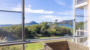 Una habitación con un amplio ventanal que da a un jardín exuberante y una masa de agua con una montaña al fondo.