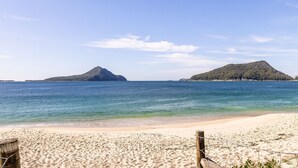 Una playa con agua azul cristalina, orilla arenosa y dos colinas destacadas al fondo.