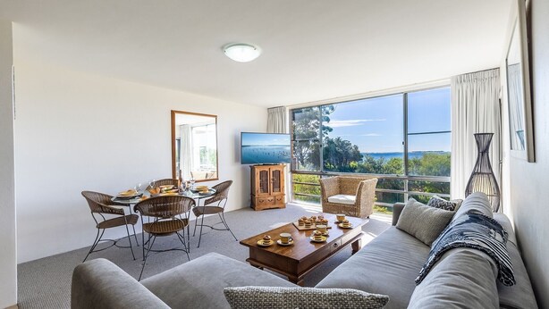 Una sala moderna con un gran ventanal que ofrece una vista panorámica, una zona de comedor con sillas de mimbre y una acogedora sala de estar con sofá y cojines.