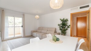 Una sala de estar moderna con un sofá color beige, una mesa de comedor blanca con un mantel tejido y una planta grande en maceta.