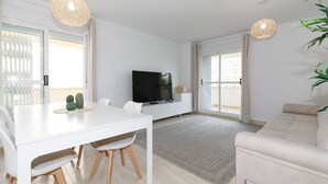Una sala de estar moderna con un sofá blanco, una mesa de comedor con sillas, un televisor de pantalla plana y un amplio ventanal con cortinas.