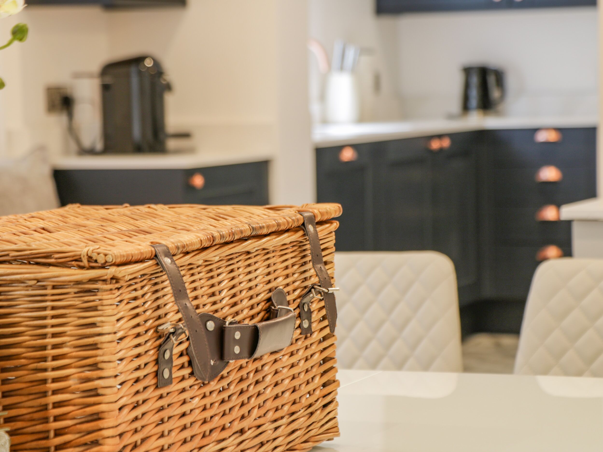 Un panier en osier avec des lanières en cuir, posé sur une table dans une cuisine moderne aux armoires blanches et au comptoir blanc.