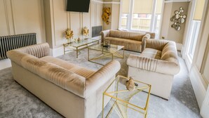 Een woonkamer met beige banken, een glazen salontafel en een aan de muur bevestigde televisie.
