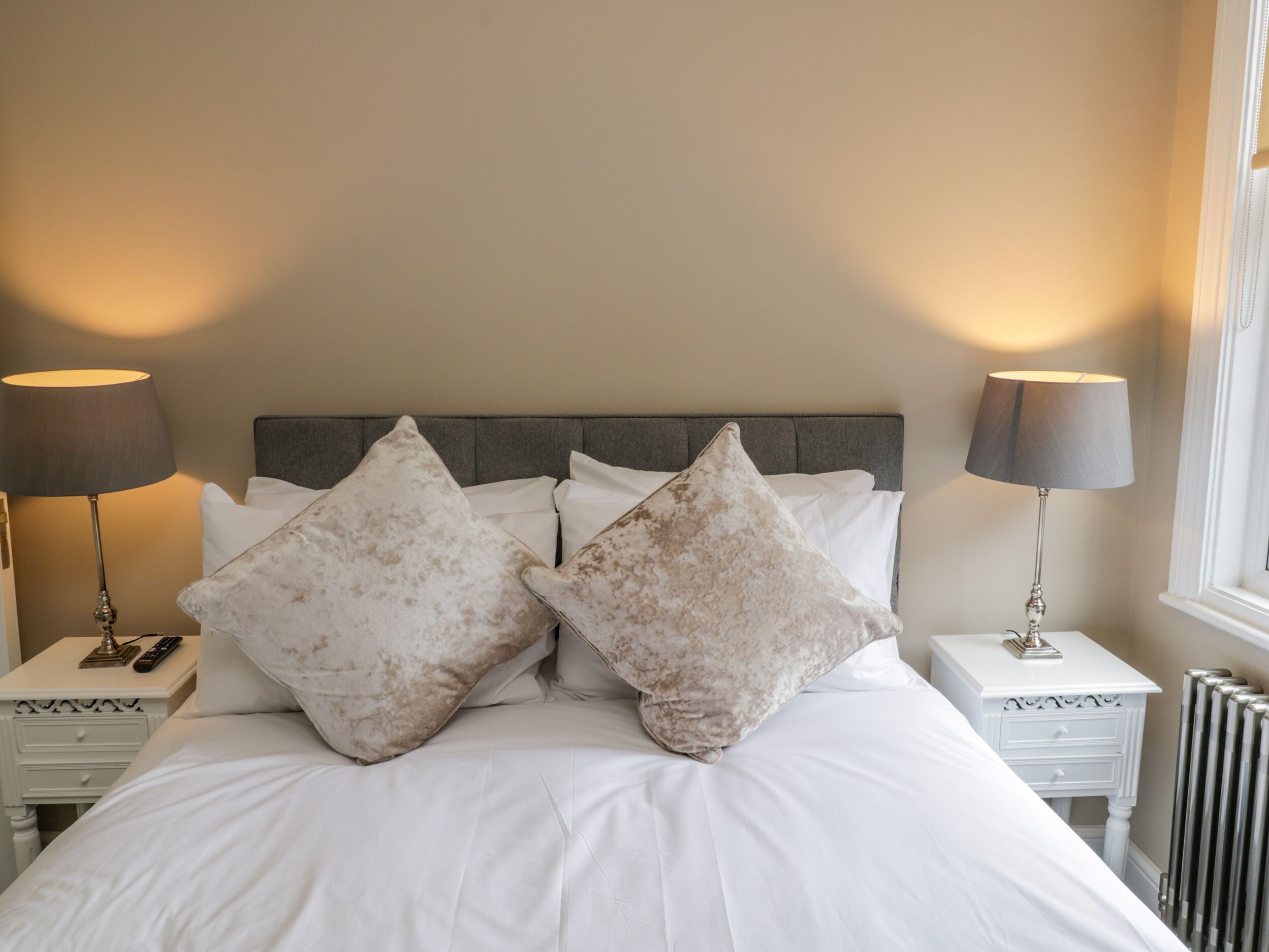 A neatly made bed with white linens and two large, light-colored pillows. There are two bedside tables with lamps and a small drawer on each. The room has a simple, clean design with a neutral color palette.