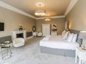 Quarto com uma cama grande, um espaço de estar com uma poltrona e uma mesa pequena, lareira e um espelho na parede.
