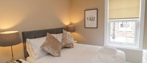 A neatly made bed with white linens and two decorative pillows, a bedside table with a lamp and remote control, and a framed sign on the wall.