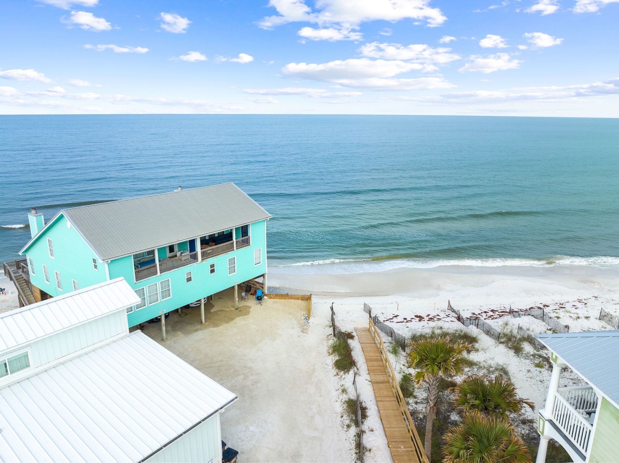 A turquoise two-story beach house with a balcony overlooking the ocean, a sandy beach, and a wooden boardwalk.