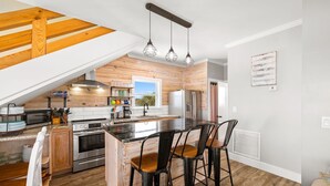 A modern kitchen with a wooden ceiling, a central island with bar stools, stainless steel appliances, and a view of the outdoors. - Gulf Front | Outdoor Shower | Dogs Allowed (Port St. Joe)