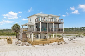 A two-story beach house with a wooden deck, a covered patio, and a view of the beach.