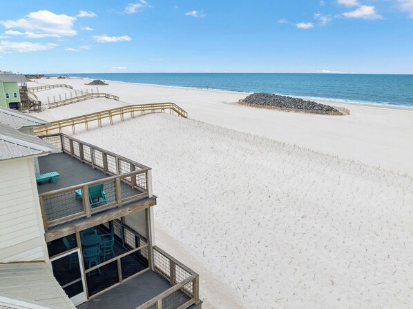 A beach view with a wooden deck, a building with a balcony, and a clear sky.