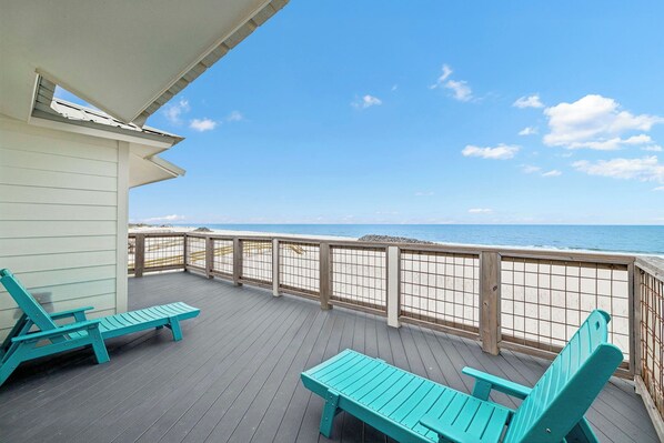 A wooden deck with two turquoise lounge chairs and a wooden railing overlooking a beach.