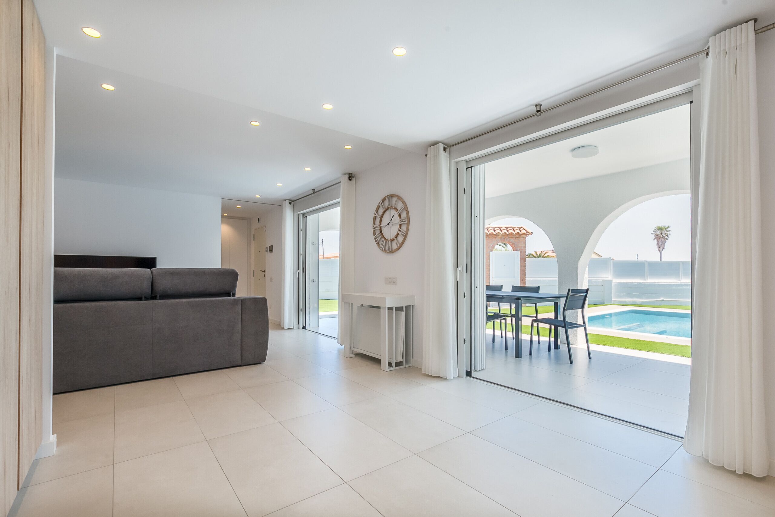 Una sala de estar moderna con un sofá gris, una mesa de comedor con sillas, y vista a una piscina a través de puertas de cristal grandes.