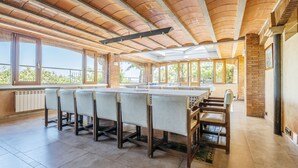 Un'ampia stanza con un lungo tavolo da pranzo e sedie, grandi finestre e un soffitto alto con travi a vista.