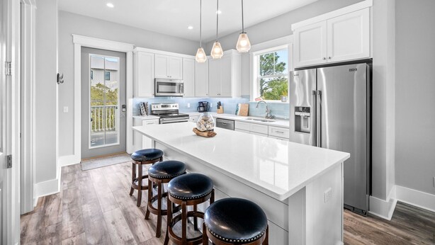 A modern kitchen with white cabinets, stainless steel appliances, a large island with stools, pendant lighting, and a view to the outdoors.