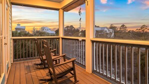 A wooden deck with a railing, two rocking chairs, and a view of a sunset over a residential area.