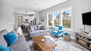 A modern living room with a grey sofa, blue armchair, and wooden coffee table. There is a TV on the right and a dining area in the background.