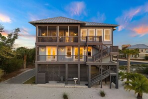 A multi-story beachfront property with balconies, a staircase, and a sandy driveway.