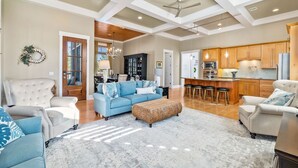 Un salon spacieux avec un canapé bleu, un fauteuil blanc et un grand pouf en osier. On aperçoit au fond une cuisine avec des meubles en bois et un coin bar.