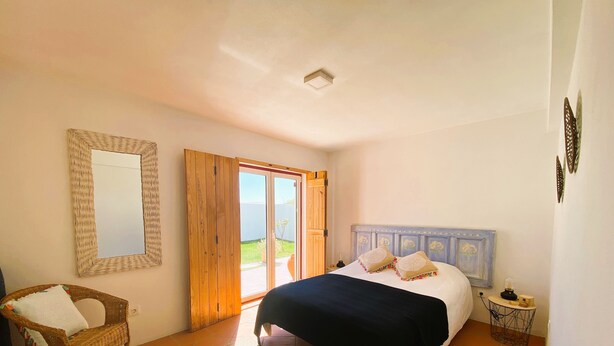 Ein Schlafzimmer mit einem Bett, einem Korbsessel, einem großen Spiegel und Blick ins Freie durch eine Schiebetür.