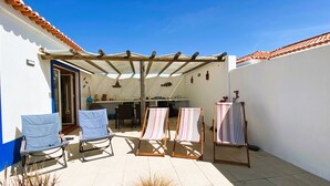 Eine Terrasse mit einer Pergola, zwei Liegestühlen, einem Tisch und einer kleinen Küchenzeile.