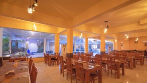 Un vaste espace dédié à la salle à manger, avec de nombreuses tables et chaises en bois, de grandes fenêtres donnant sur l’extérieur et des luminaires suspendus.