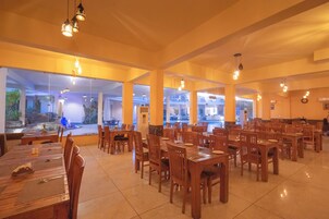 A spacious dining area with multiple wooden tables and chairs, large windows offering views of the outdoors, and hanging pendant lights.