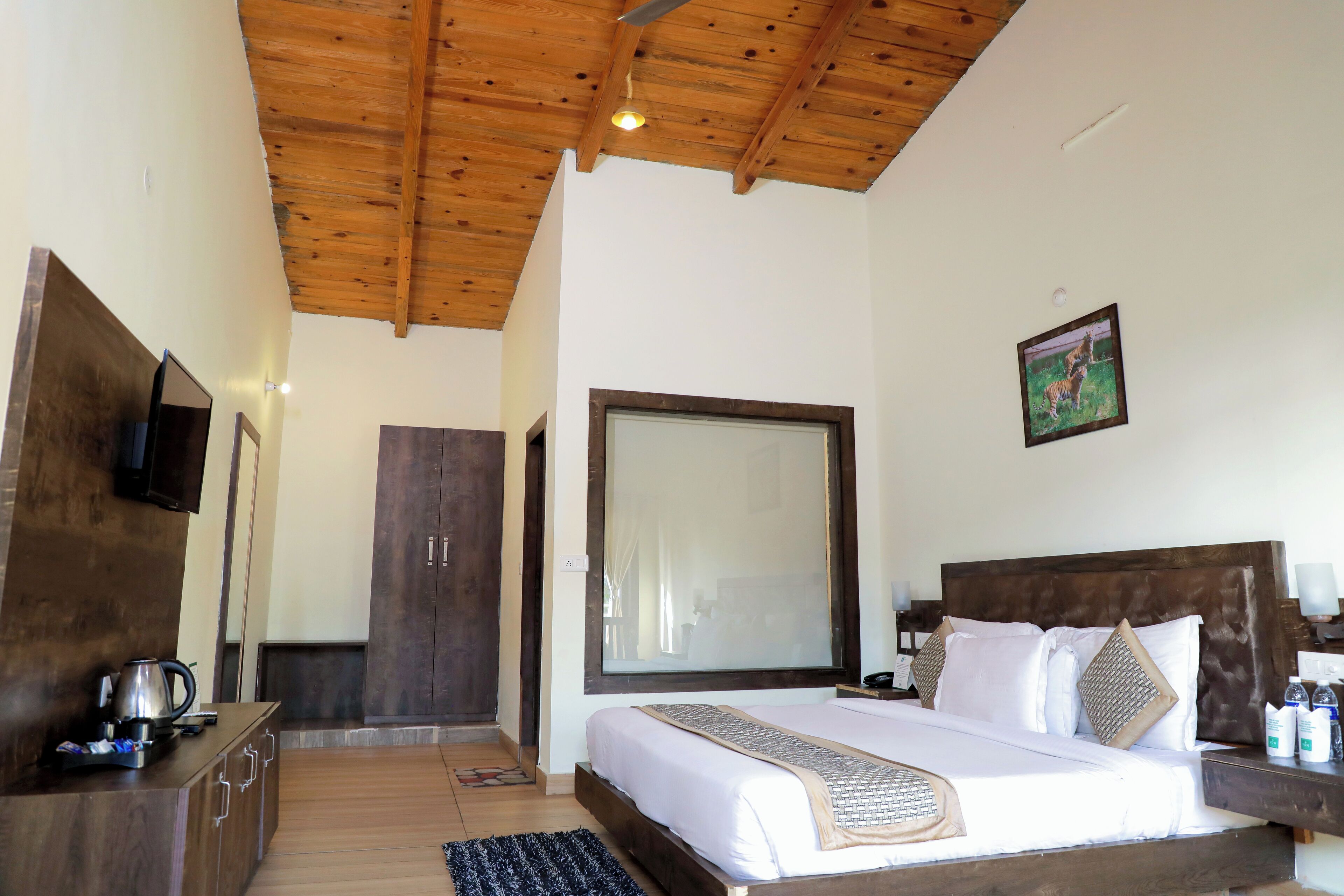 Un dormitorio con techo de madera, un ventanal grande, una cama con ropa de cama blanca, un aparador de madera y un televisor en la pared.