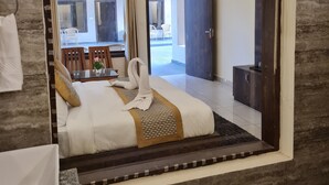 Une chambre d’hôtel dotée d’un grand miroir, d’un lit recouvert d’une couvre-lit blanche et dorée, d’une tête de lit en bois, d’une salle de bain avec un lavabo et un miroir, et offrant une vue sur l’intérieur de la pièce.