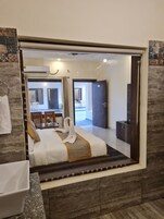Habitación de hotel con un espejo grande, cama con cubrecama blanco y dorado, cabecero de madera, baño con lavamanos y espejo, y vista al interior de la habitación.