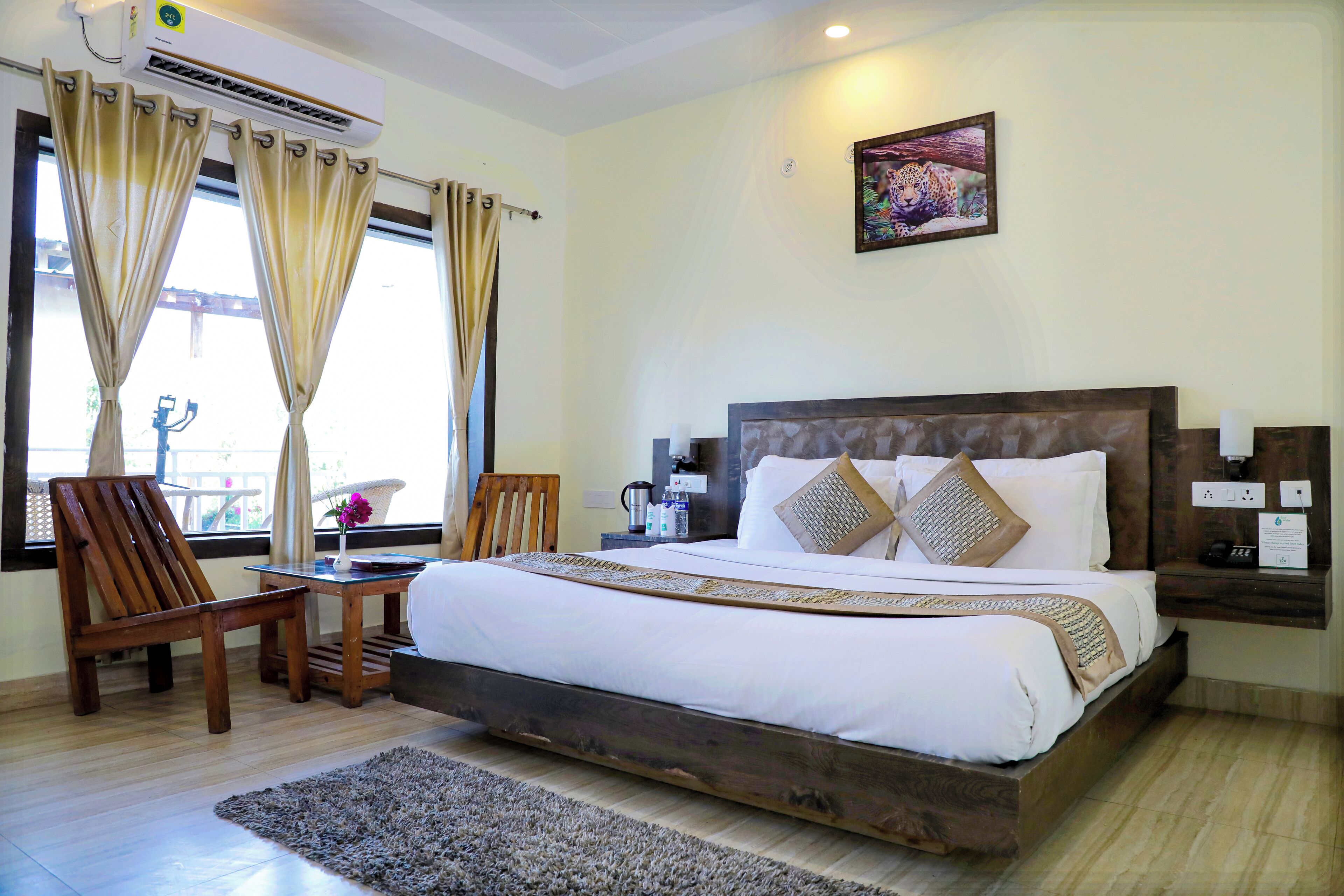 A hotel room with a large bed, wooden headboard, two chairs, a small table, and a window with curtains.
