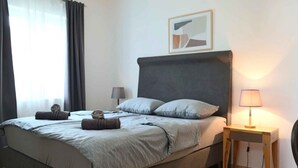 En pænt redt seng med et gråt sengegavl, et natbord med en lampe, og et indrammet billede på væggen.