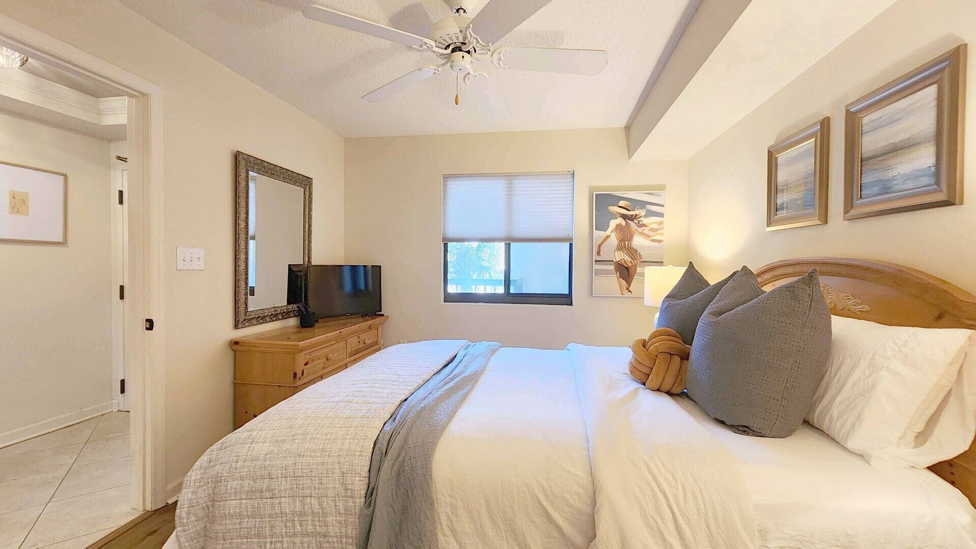 Une chambre à coucher avec un lit, un téléviseur posé sur une commode en bois, un ventilateur de plafond, une fenêtre et des œuvres d’art encadrées accrochées au mur.