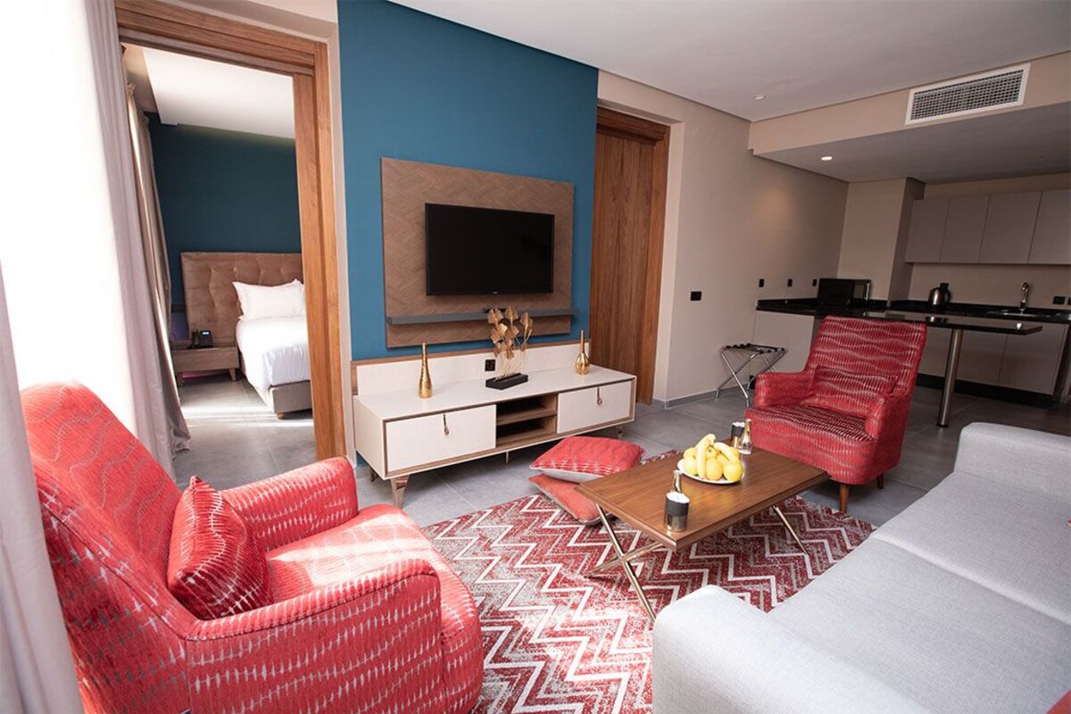 A modern hotel room with a bed, a TV mounted on the wall, a dining area, and a seating area with red chairs and a white sofa.