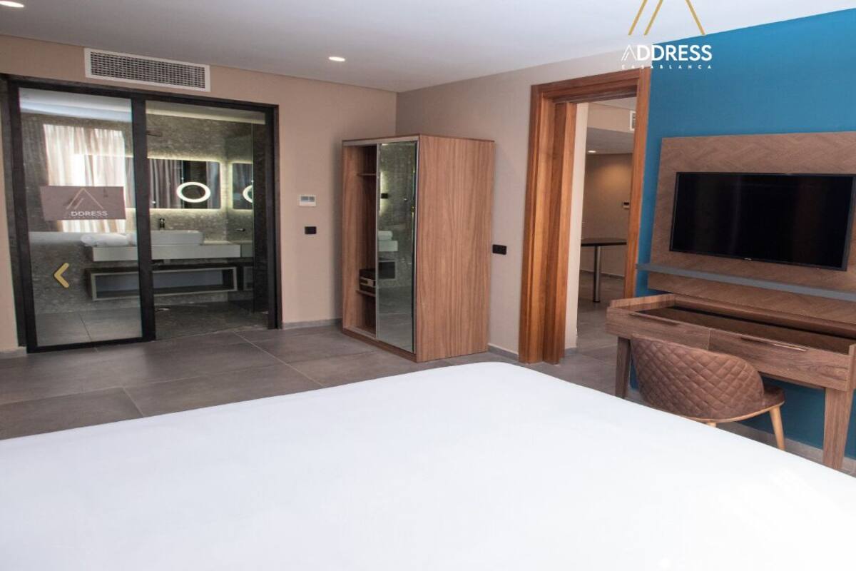 Una habitación de hotel moderna con una cama grande, un televisor montado en la pared, un escritorio y una zona de estar con una silla roja.