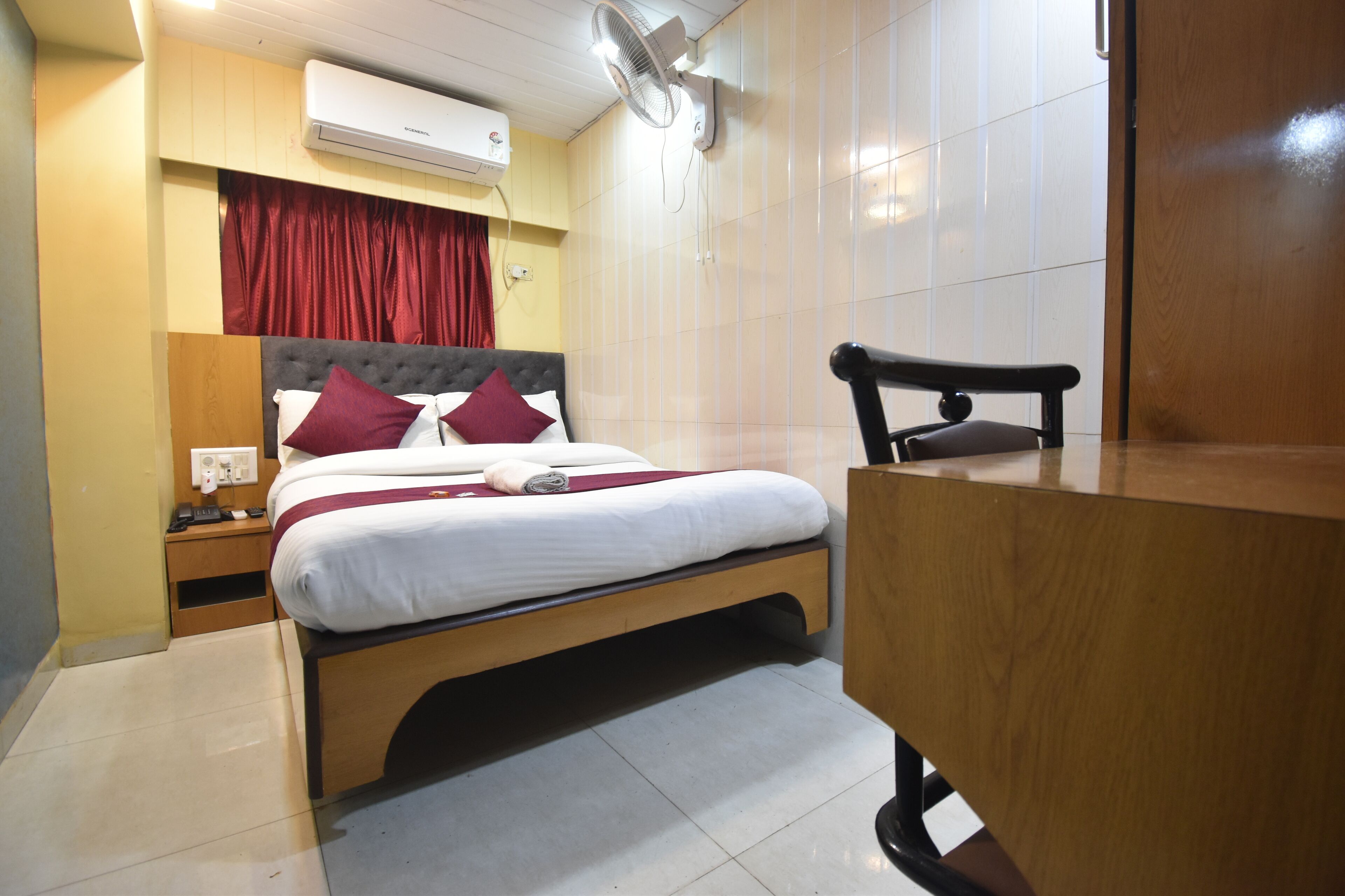 Una habitación de hotel con cama, mesita de noche, escritorio, silla, aire acondicionado y ventilador de pared.