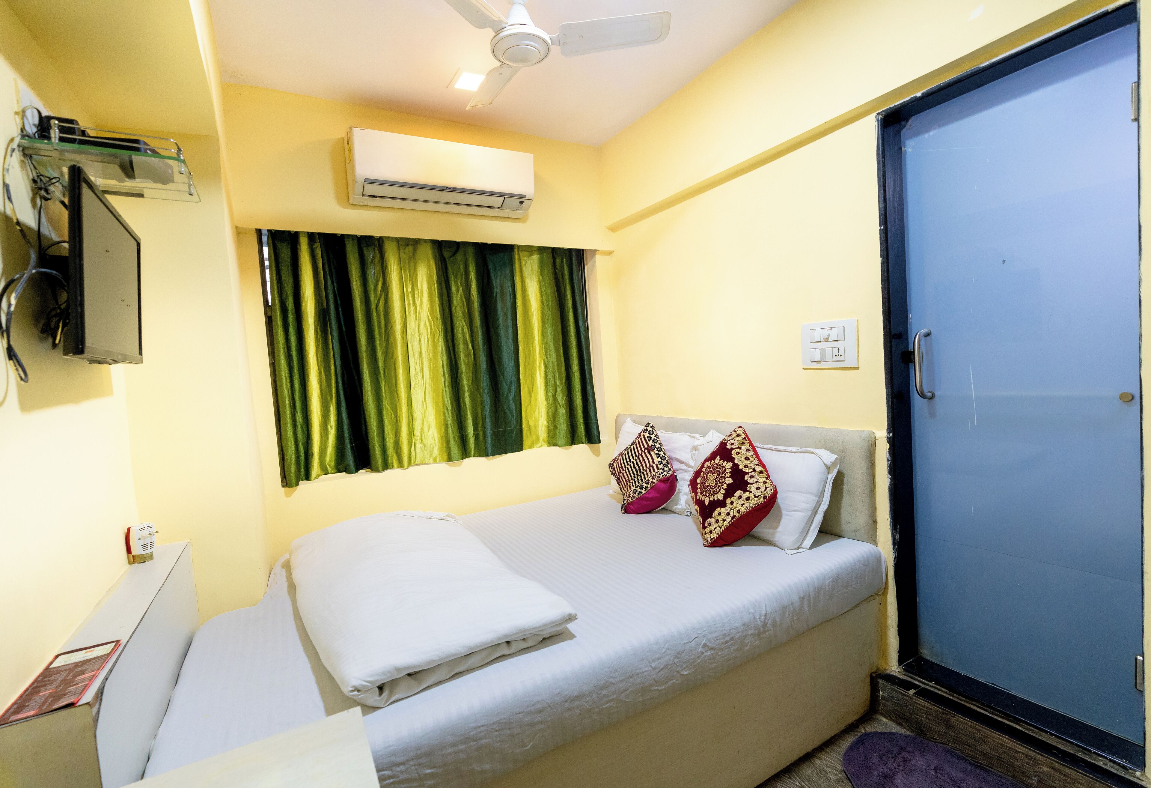 Habitación de hotel con cama, unidad de aire acondicionado, ventilador de techo, puerta y ventana con cortinas.