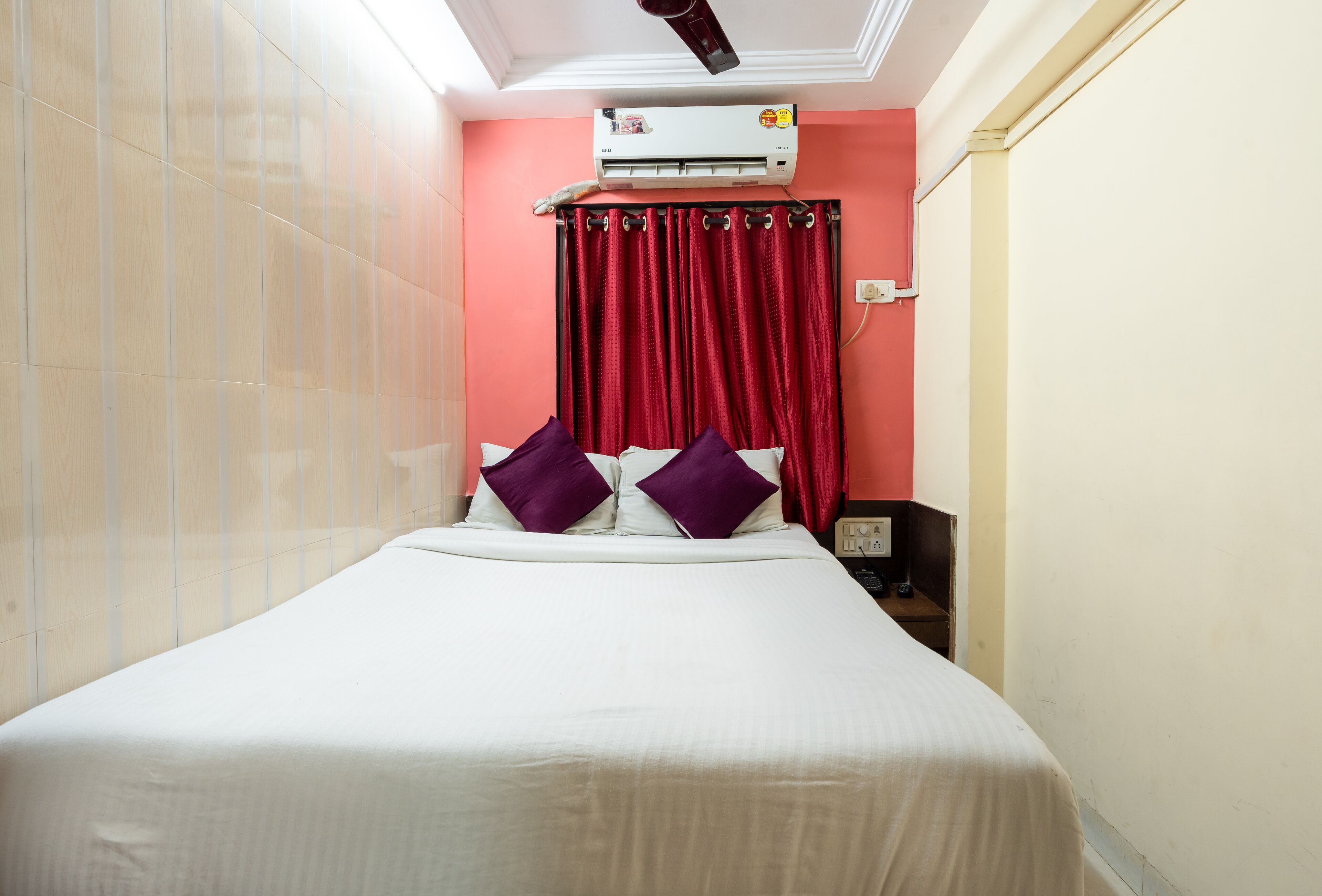 Una habitación de hotel con cama, cortinas rojas, equipo de aire acondicionado y mesita de noche.