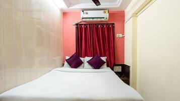 Una habitación de hotel con cama, cortinas rojas, equipo de aire acondicionado y mesita de noche.