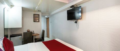 A hotel room with a bed, a desk, a chair, a television mounted on the wall, and a small refrigerator.