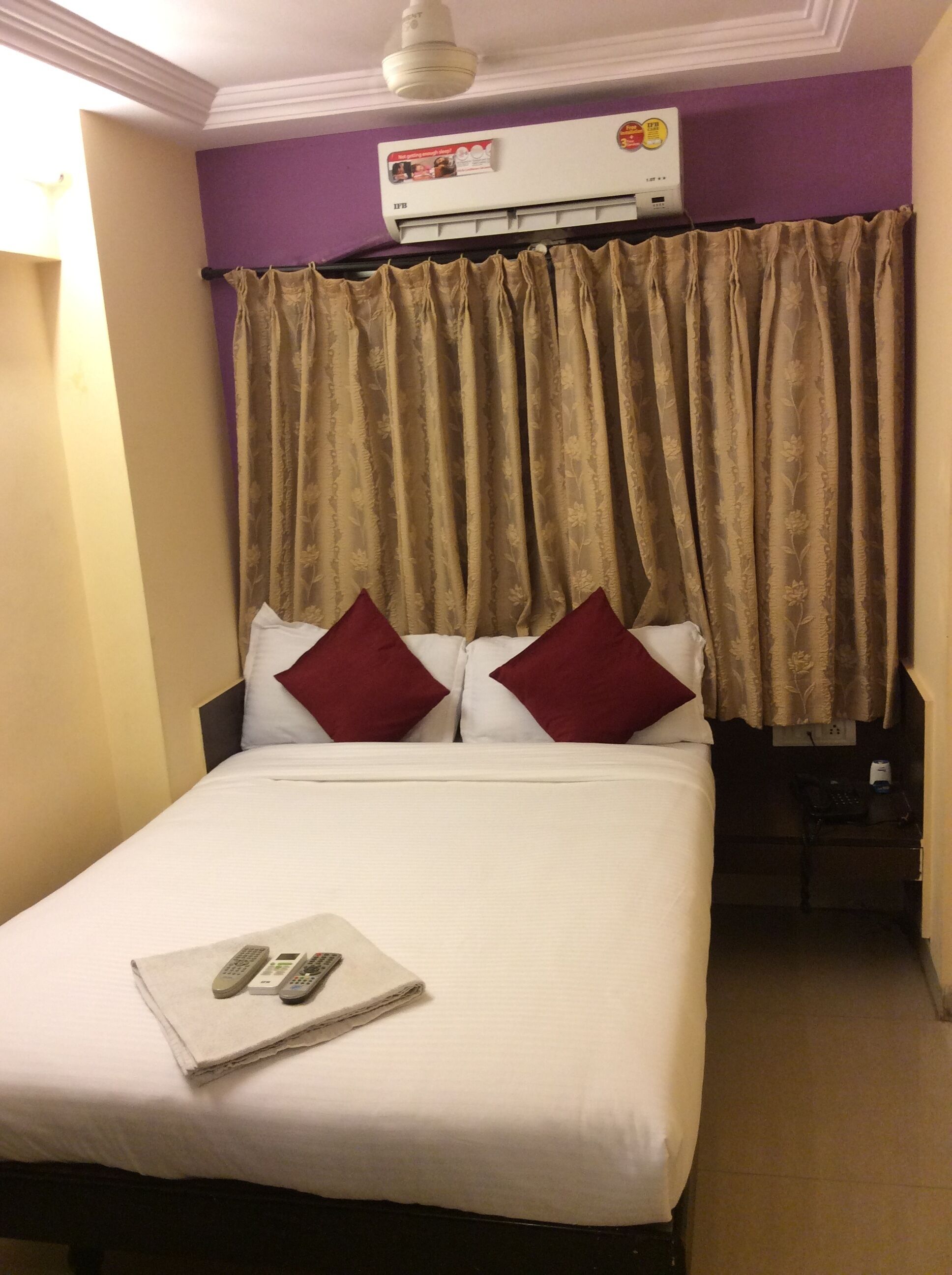 A hotel room with a bed, two red pillows, a white blanket, and a remote control on the bed.