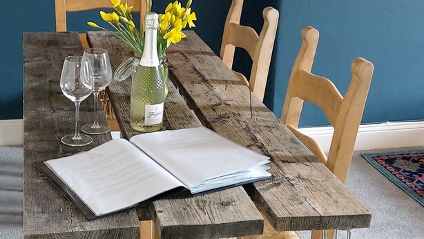 Ein rustikaler Holztisch mit einer Vase voller gelber Blumen, zwei Weingläsern und einem offenen Buch.