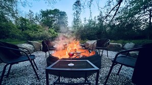 Een vuurkorf met stoelen eromheen, op een grindpad, met bomen op de achtergrond.