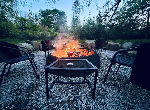 Ein Lagerfeuer mit umliegenden Stühlen, auf einer Schotterfläche, mit Bäumen im Hintergrund.