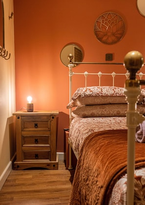 A bedroom with a wooden nightstand, a bedside table, a bed with a floral-patterned bedspread, a round mirror on the wall, and a wall-mounted light fixture.