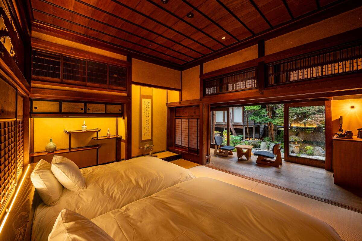 A traditional Japanese-style room with tatami flooring, a large bed, sliding doors leading to a garden, and a view of the surrounding nature.