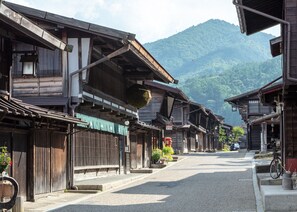 A traditional wooden street with buildings, a bicycle, and a mountain in the background. - BYAKU Narai (Shiojiri)